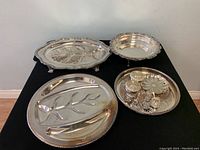 Overall view of various vintage silver plated serving trays including the largest footed oval tray, round platter, divided tray with deer motif, and the tray with multiple small containers and leaf spoons.