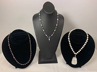 Three vintage pearl necklaces displayed on black busts: a white baroque pearl choker with abalone pendant, a black/peacock pearl necklace, and a blue/white pearl drop necklace.