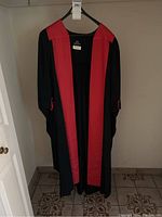 Full view of black ceremonial robe with red trim hanging from hanger against wall.