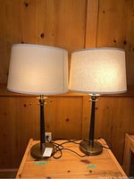 Pair of table lamps shown side by side on a wooden surface against a wood-paneled wall, each with a white fabric shade and black and brass base.