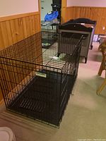Full view of black collapsible wire dog cage with plastic tray on floor in room with wood panel walls.