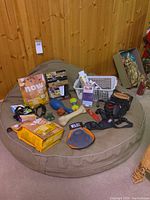 Overview of all items placed on large beige dog bed, showing dog food, chew toys, halter, leash, grooming glove, and training system box