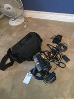 Photo showing the Sony Handycam video camera, black carrying bag, charger, battery pack, and AV cables laid out on carpet.