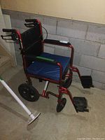 Side angle of red transfer chair showing black padded backrest, blue cushioned seat, footrests, wheels, and hand brakes