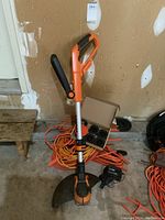 Worx cordless string trimmer leaning against wall with extension cords coiled underneath. Charger and box of spools visible.