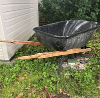 Side angle view of the black metal wheelbarrow with wood handles and flat tire in a grassy outdoor setting beside a building