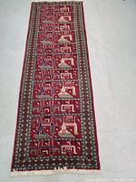 Top view of the full Persian rug runner showing deep red color with geometric and architectural pattern blocks in a repeating sequence, bordered with intricate diamond and floral motifs, fringed on one end.