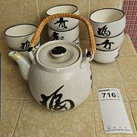 Ceramic teapot with bamboo-wrapped handle and eight stacked ceramic cups on tiled surface.