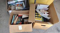 Photo shows three large cardboard boxes filled with vintage books and paper materials, some books are upright and others stacked horizontally.