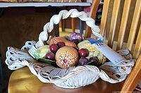 Front view of the Capodimonte ceramic fruit basket centerpiece placed on a chair showing woven basket with handle and various ceramic fruits inside.