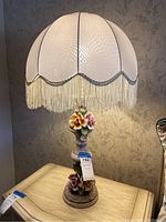 Full view of the Capodimonte porcelain table lamp on a wooden table, showing the fabric frill shade with fringe and floral porcelain base.