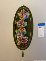 Full view of oval green fabric plaque with colorful porcelain flowers and gold trim, with seller's lot tag attached.