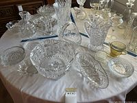 Full display of assorted crystal and pressed glass tableware on round white cloth-covered table, showing various bowls, vases, pitchers, and dishes in intricate cut patterns.