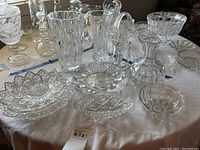 Full assortment of glass and crystal tableware including bowls, vases, and decorative dishes arranged on a white tablecloth
