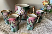 Porcelain pitcher and four mugs grouped together on a cloth surface, showing floral grapevine designs and gold rim accents.
