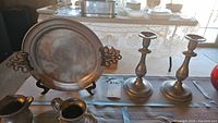 Photo showing the pewter plate on a black stand with ornate side handles, two pewter candlesticks, and a pewter creamer on a table.