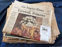 Stacked vintage newspapers with Los Angeles Times front page headline 'Farewell, Princess' and a photo of Princess Diana's funeral coffin draped with flowers.