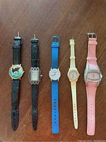 Full view of 5 novelty and fashion wristwatches laid flat with visible dials and straps to show variety of designs and materials.