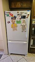 Front view of a white Whirlpool Gold fridge with a bottom freezer, decorated with magnets and paper cutouts, showing the overall appliance condition and size in kitchen context.