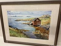 Full view of the framed original watercolour painting showing coastal landscape with red boathouse on stilts over water and rocky shore.