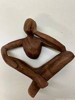 Front view of the wooden carved figure seated cross-legged with a hand on the face.