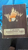 Front cover of the vintage 1929 Art Deco style cookbook titled 'The Modern Housekeepers' Guide' with special reference to gas cookery showing visible creases, wear and tear consistent with light age use.