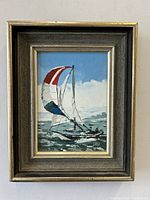 Framed oil painting showing a yacht with multi-colored sails on the ocean, signed by Nancy Leon at bottom right corner.