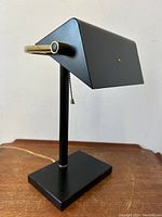 Black metal banker’s desk lamp shown unlit on wooden surface, displaying its angular shade, brass detail, and pull chain switch.