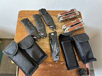 Overview of six tools on a wooden surface with three black knives, two silver multi-tools with wood handles, and one black utility knife, including five black nylon carrying pouches.