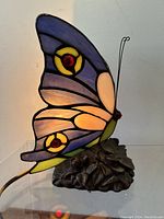 Side view of the stained glass butterfly lamp showing illuminated wings with purple, blue, and yellow glass accented with red glass pieces, mounted on a flower petal base.