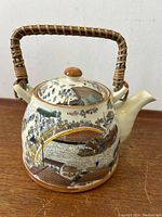 Clear front view of teapot on wooden surface showing the main painted ribbon around the body and the wicker handle.