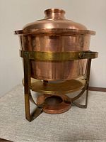Copper pot in brass warming stand with lid on top, shown on table.
