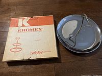 Photo showing the Kromex two-tier serving tray with original box labeled 'Holiday Giftware' and the Rivera triple chrome single level tray.