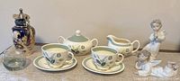 Full tea set including two Lladro angel figurines, two cups and saucers, sugar bowl, creamer, silver plated pitcher, and green glass tea light holder.