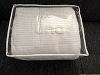 Photo of the folded Luna weighted blanket inside a clear plastic zippered storage bag, showing the white fabric with grey stripes and Luna logo faintly visible.