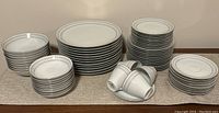 Photo showing multiple stacks of dinnerware including large dinner plates, salad plates, bowls, cups, and saucers. Items are white with gray geometric patterns around rims.