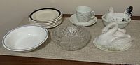 Overview of the lot showing all items arranged on a table: 5 white plates with blue rims, a Corelle bowl, a clear glass candy dish, a milk glass swan, a milk glass chicken lidded bowl, and a vintage teacup and saucer.