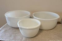 Three milk glass mixing bowls displayed together on a cloth surface. Two large bowls and one small bowl, all white opaque milk glass with smooth surfaces.