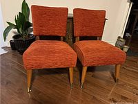 Front view showing the pair of orange vintage chairs with wooden tapered legs and brass leg caps, orange fabric upholstery with textured weave.