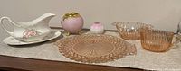 Setup on a cloth with all items visible: pink depression glass plate and bowl, pink and gold footed vase, floral porcelain gravy boat with saucer, small pink and white vase.