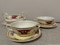 Side view of gravy boat with matching oval saucer and two two-handled soup bowls on their saucers with burgundy and floral designs.