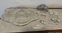 Photo showing glass footed cake plate with silver inlay, metal napkin holder, and silver plate flatware arranged on table