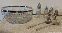 Clear cut glass bowl with silver rim, next to silver-plated salad spoon and fork, glass and silver salt and pepper shakers, and glass sugar shaker