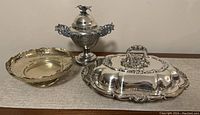 Wide shot showing all three items: the covered entree dish with ornate lid and handle on the right, the sugar bowl centerpiece with finial in the middle, and the round footed scalloped dish on the left.