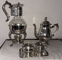 Overall photo showing the complete coffee service set including the glass and silver plated coffee pot, silver plated teapot, cream and sugar set on a silver tray.