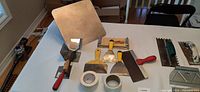 Photo showing assorted drywall mudding and finishing tools including trowels, putty knives, corner finishing tools, and partial rolls of masking tape all laid out on a table.