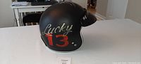 Side view of matte black motorcycle helmet with Lucky 13 painted on the side in gold and red.