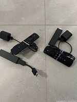 Two Amazon Fire TV Sticks with remotes and power adapters laid on a tiled floor, cables wrapped with rubber bands.