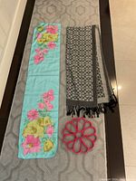Three items laid flat: pink felt scarf holder on bottom, grey patterned scarf with black fringes on right, light blue floral scarf on left.
