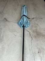 Blue outdoor umbrella closed, showing black metal pole and folded blue fabric canopy on concrete floor.